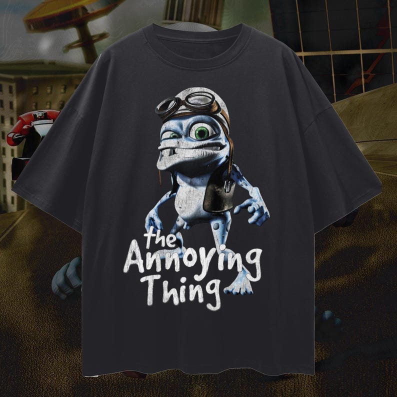 May include: Dark gray t-shirt featuring a cartoon frog character wearing a pilot's helmet and goggles. The frog is blue and white with green eyes. The text "the Annoying Thing" is written in white script below the frog.