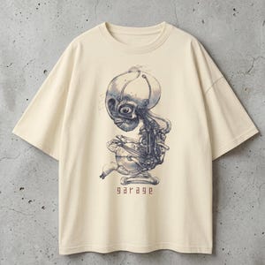 May include: Cream-colored t-shirt featuring a detailed, blue and gray illustration of a mechanical creature. The design includes intricate details and the word "garage" in a stylized font at the bottom. The shirt has a relaxed fit.