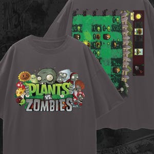 Puede incluir: Camisetas de color gris oscuro con el juego Plants vs. Zombies. La parte delantera de la camiseta muestra el logotipo del juego y los personajes, incluyendo un girasol, un zombi y varias plantas. La parte trasera muestra un nivel del juego.