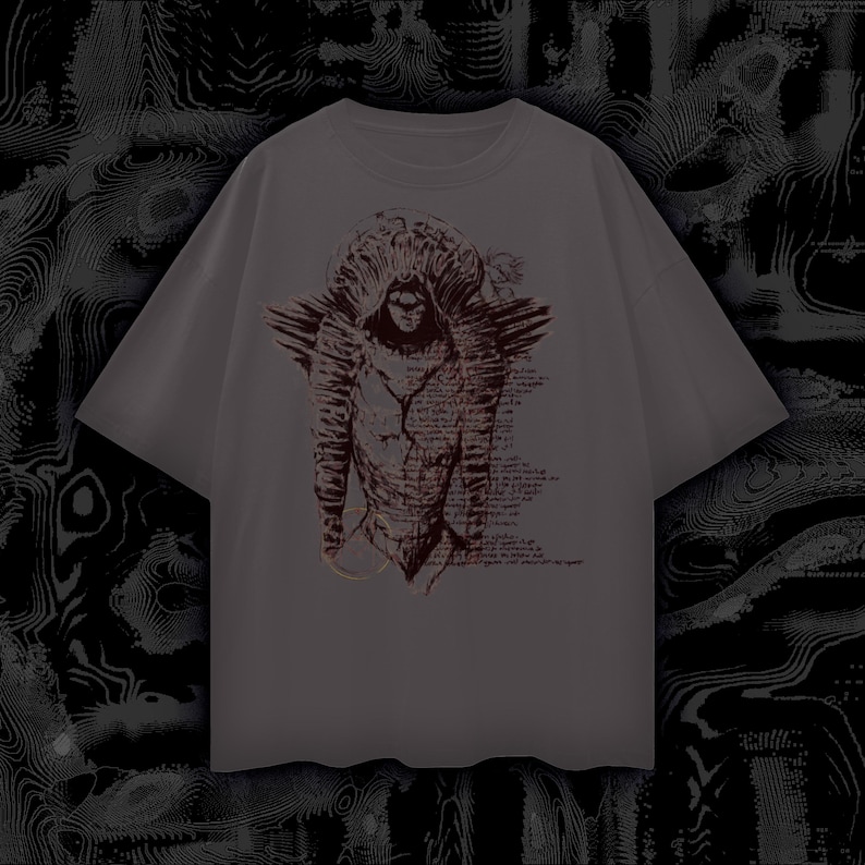 May include: Grey t-shirt with a detailed illustration of a figure with a horned headdress and wings. The artwork is in shades of brown and red, with script-like text incorporated into the design. The shirt has short sleeves and a relaxed fit.