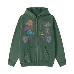 Pode incluir: Um casaco com capuz e fecho de correr em verde escuro com uma estampa gráfica frontal com formas abstratas coloridas, padrões florais e motivos circulares escuros em ambos os lados do fecho. Inclui capuz, mangas compridas e um bolso frontal.