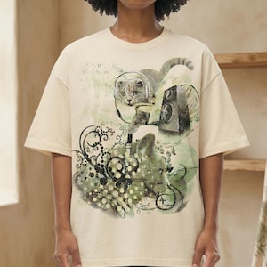 May include: A beige short-sleeve t-shirt featuring a graphic print of a cat wearing headphones, a speaker, and abstract swirling patterns in shades of green and black. The design includes circular motifs and intricate line work across the front.