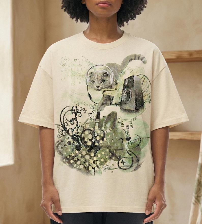 May include: A beige short-sleeve t-shirt featuring a graphic print of a cat wearing headphones, a speaker, and abstract swirling patterns in shades of green and black. The design includes circular motifs and intricate line work across the front.