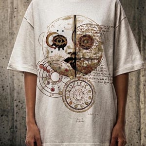 Pode incluir: Camiseta cinza claro oversized com um gr&aacute;fico detalhado de um design celestial. O design inclui um rosto de meia-lua, engrenagens e elementos circulares com escrita. A camisa tem mangas curtas e gola redonda.