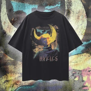 May include: Black t-shirt featuring a surreal graphic of a figure with a yellow crescent-shaped headpiece and a blue turtleneck. The text "HYFI-CS" is printed below the figure in a stylized font. The background has a colorful, abstract design.