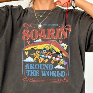 May include: Dark gray t-shirt featuring a colorful graphic of Mickey Mouse, Donald Duck, Goofy, and Pluto hang gliding. The text on the shirt reads "Commander Soarin' Around The World" in a vintage style.