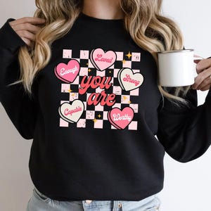 May include: Black sweatshirt with a checkerboard heart design. The design features pink hearts with the words "Enough", "Loved", "Strong", "Capable", and "Worthy". The words "you are" are in a red script font. A white mug is held in the hand.