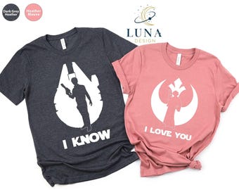 May include: Two short-sleeved t-shirts, one dark gray and one mauve. The dark gray shirt features a white graphic of a spaceship and a figure with the text "I KNOW". The mauve shirt has a white graphic of a symbol and figure with the text "I LOVE YOU".