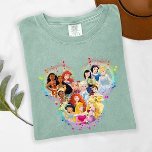 May include: A light green Comfort Colors t-shirt featuring a heart-shaped graphic of Disney princesses. The graphic includes characters like Ariel, Snow White, and Cinderella, surrounded by colorful stars. The shirt is a size L-G.