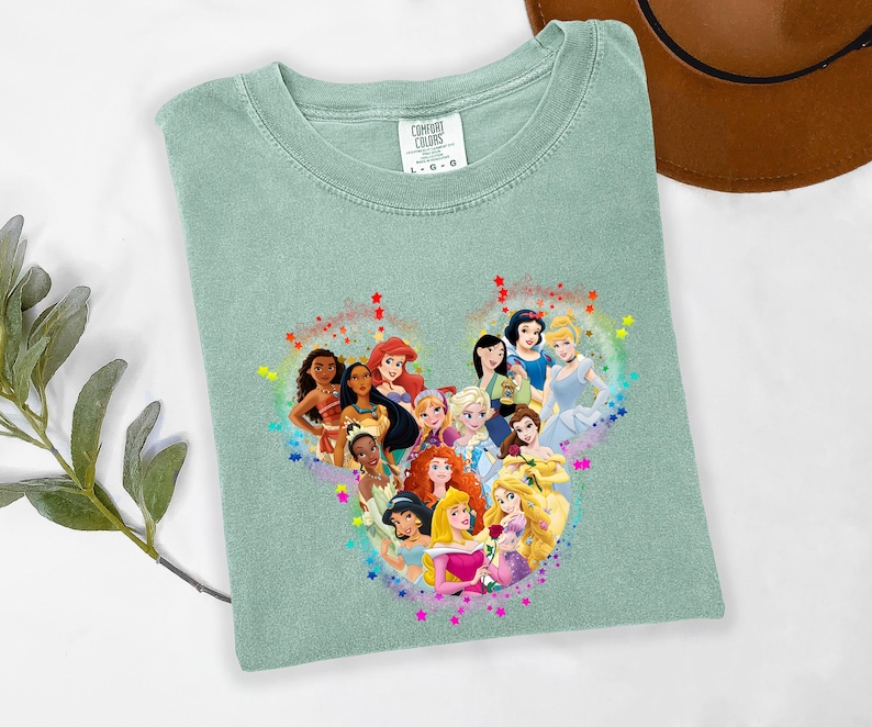 May include: A light green Comfort Colors t-shirt featuring a heart-shaped graphic of Disney princesses. The graphic includes characters like Ariel, Snow White, and Cinderella, surrounded by colorful stars. The shirt is a size L-G.