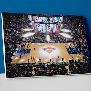 May include: A canvas print of Madison Square Garden during a New York Knicks basketball game. The court is visible with the Knicks logo at center court. The scoreboard displays "NEW YORK" in large letters. The stands are filled with spectators.