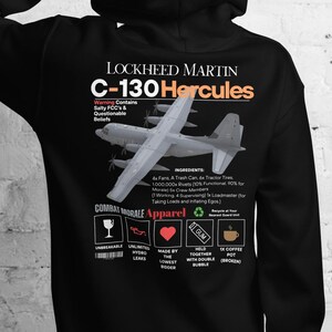 Könnte beinhalten: Schwarzer Kapuzenpullover mit einer Grafik eines Lockheed Martin C-130 Hercules Flugzeugs. Das Design enthält den Text "Lockheed Martin C-130 Hercules" in Weiß und Orange. Zusätzlicher Text und Symbole sind ebenfalls auf der Rückseite vorhanden.