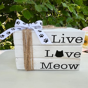 以下が含まれることがあります： 「Live Love Meow」の黒い文字が書かれた、3つの白い木製ブロックのスタック。黒い足跡模様の白いリボンが一番上のブロックに結ばれています。ジュートの紐がブロックに巻き付けられています。ピンクと黄色の花が背景にあります。