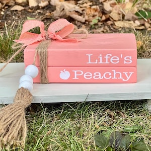 Op de afbeelding: Een decoratieve stapel van twee roze houten boeken met de tekst "Life's Peachy" in het wit. De boeken zijn vastgebonden met touw en een roze lint en liggen op een wit houten dienblad met een kwastje en kralenaccent.