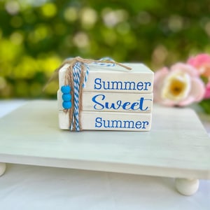 May include: A stack of three white wooden blocks with the words "Summer Sweet Summer" in blue script. The blocks are tied with twine and blue and white string, and adorned with blue beads. The blocks sit on a white, square, footed tray.