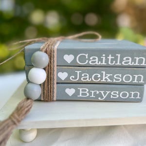 May include: A stack of three gray decorative books tied with twine, featuring the names "Caitlan", "Jackson", and "Bryson" in white, each with a heart symbol. A tassel and decorative beads hang from the twine.