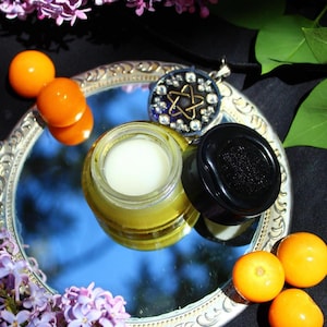 Peut inclure: Un miroir argenté reflète un ciel bleu, présentant un petit pot de crème, un pot noir et un pendentif avec un motif d'étoile. Des baies oranges et des fleurs violettes entourent le miroir, créant une esthétique naturelle et mystique.