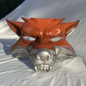 Peut inclure: Un masque de renard orange et argenté fait à la main. Le masque présente des oreilles pointues, un museau détaillé et des dents pointues. Les yeux sont ouverts et il y a des accents argentés au-dessus des yeux. Le masque est probablement destiné à un costume ou à des fins décoratives.