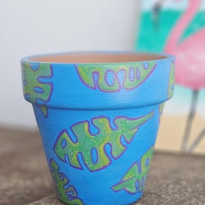 Puede incluir: Una maceta de terracota azul con un diseño pintado a mano de hojas tropicales verdes y moradas.