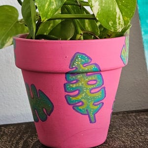 Puede incluir: Una maceta de terracota rosa con hojas pintadas de verde y amarillo. La maceta está llena de una planta verde y amarilla variegada.