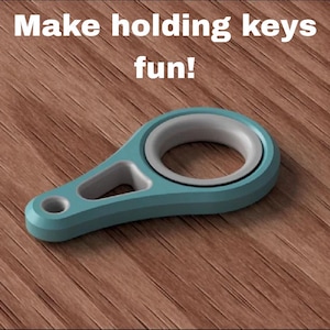 May include: A teal key holder with a gray interior ring and a keyhole. The text "Make holding keys fun!" is displayed in white above the key holder. The key holder is on a brown wooden surface.
