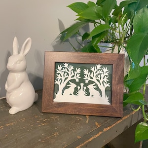 May include: A framed paper cut-out art piece with a dark brown frame. The art features two white rabbits silhouetted against a green background with trees and flowers.
