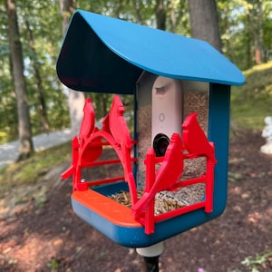 May include: A blue and orange bird feeder with red cardinal decorations. The feeder has a camera installed and is filled with birdseed. The feeder is mounted on a black pole and is outdoors.