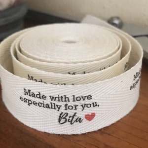 K&ouml;nnte beinhalten: Wei&szlig;es Baumwoll-Twillband mit einer schwarzen bedruckten Nachricht: "Made with love especially for you, Bita" mit einem kleinen roten Herzen.