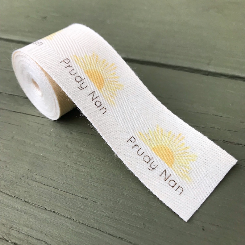 Op de afbeelding: Wit katoenen lint met een gele zonafbeelding en de tekst "Prudy Nan" gedrukt in bruin.