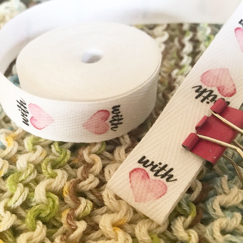 K&ouml;nnte beinhalten: Wei&szlig;es Baumwollband mit rosa Herzen und dem Wort "with" darauf gedruckt. Das Band ist aufgerollt und es gibt ein loses Ende mit einem rosa Herzen und dem Wort "with" darauf gedruckt.