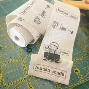Op de afbeelding: Een wit katoenen lint met de tekst "human made" in zwart gedrukt. Het lint is gevouwen en vastgeklemd met een zilveren paperclip. De tekst "Sew Every Whim" is ook op het lint gedrukt.
