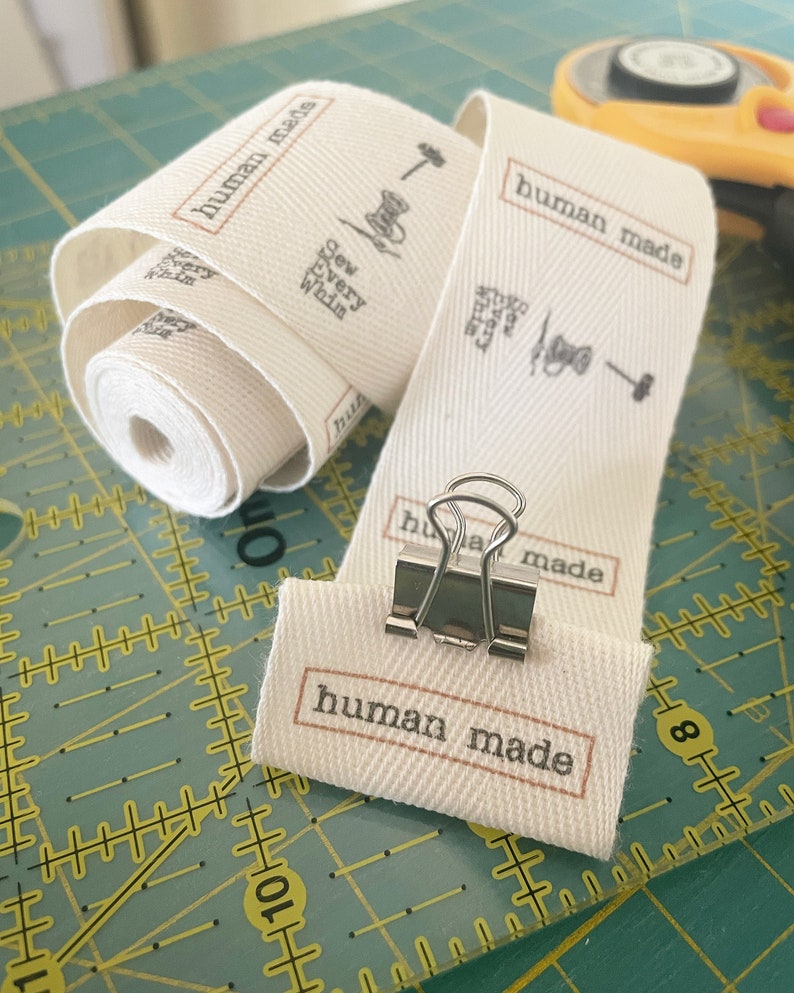 Op de afbeelding: Een wit katoenen lint met de tekst "human made" in zwart gedrukt. Het lint is gevouwen en vastgeklemd met een zilveren paperclip. De tekst "Sew Every Whim" is ook op het lint gedrukt.