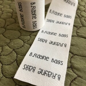 Op de afbeelding: Een wit katoenen lint met de tekst "B.Rayne Bags" die er herhaaldelijk op staat gedrukt. Het lint is opgerold en klaar om te worden gebruikt voor verpakking.