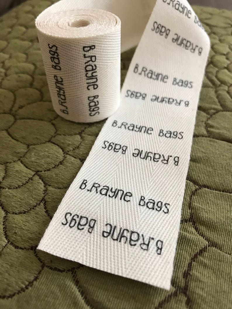 Op de afbeelding: Een wit katoenen lint met de tekst "B.Rayne Bags" die er herhaaldelijk op staat gedrukt. Het lint is opgerold en klaar om te worden gebruikt voor verpakking.