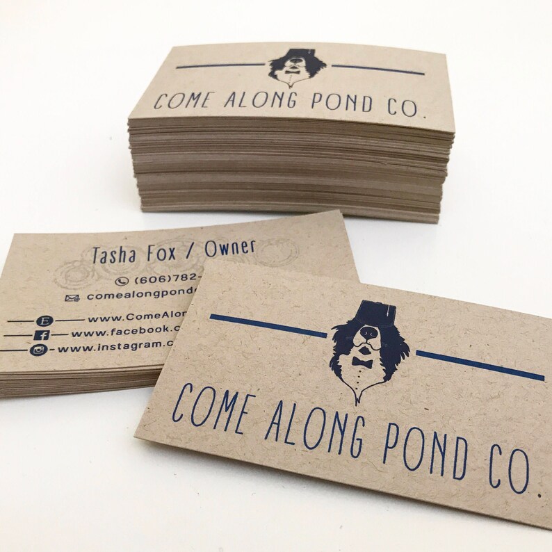 May include: A stack of brown kraft paper business cards with the text "COME ALONG POND CO." and a black and white illustration of a dog wearing a top hat and bow tie. The contact information for Tasha Fox, Owner, is listed on the card, including a phone number, email address, and links to social media.