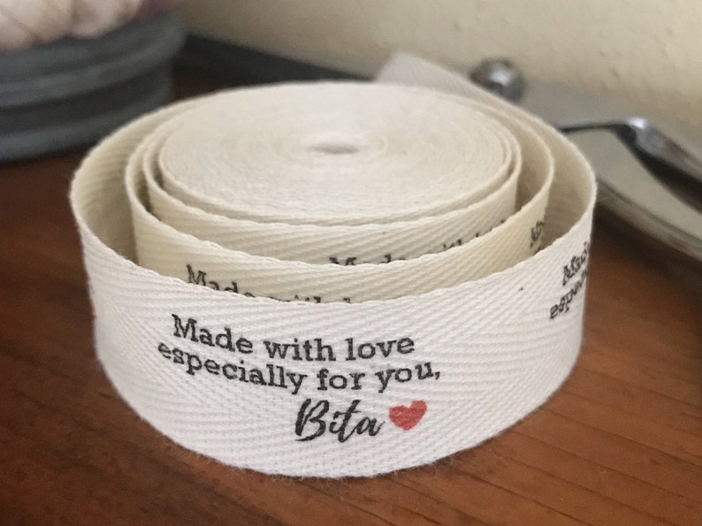K&ouml;nnte beinhalten: Wei&szlig;es Baumwoll-Twillband mit einer schwarzen bedruckten Nachricht: "Made with love especially for you, Bita" mit einem kleinen roten Herzen.