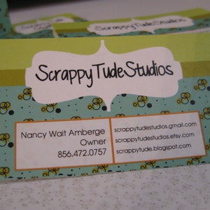 May include: A green and blue business card with a white border and a white oval with the text "Scrappy Tude Studios" in black. The card also includes the name "Nancy Wait Amberge", the title "Owner", and the phone number "856.472.0757". The card also includes the following contact information: scrappytudstudios.gmail.com, scrappytudstudios.etsy.com, and scrappytude.blogspot.com.