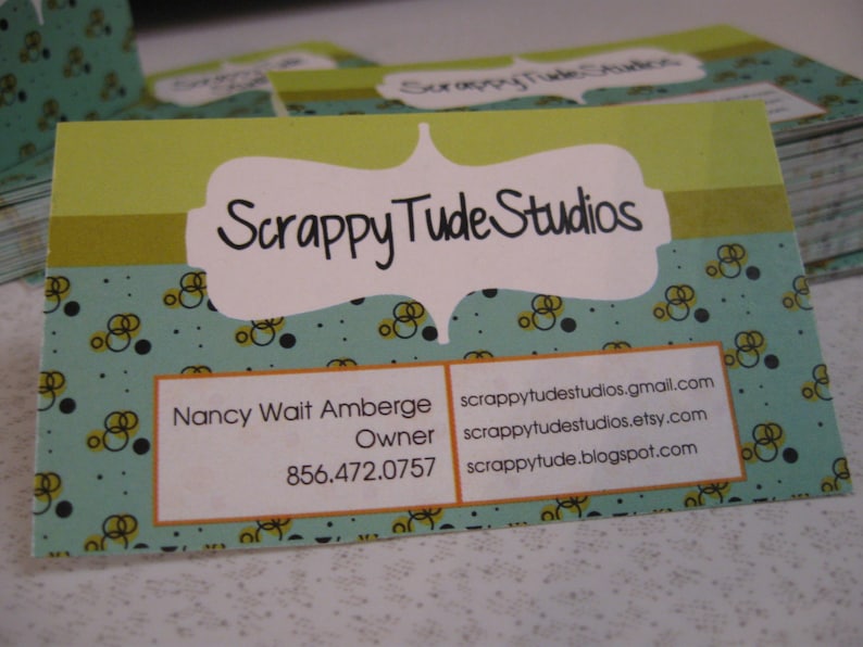 May include: A green and blue business card with a white border and a white oval with the text "Scrappy Tude Studios" in black. The card also includes the name "Nancy Wait Amberge", the title "Owner", and the phone number "856.472.0757". The card also includes the following contact information: scrappytudstudios.gmail.com, scrappytudstudios.etsy.com, and scrappytude.blogspot.com.