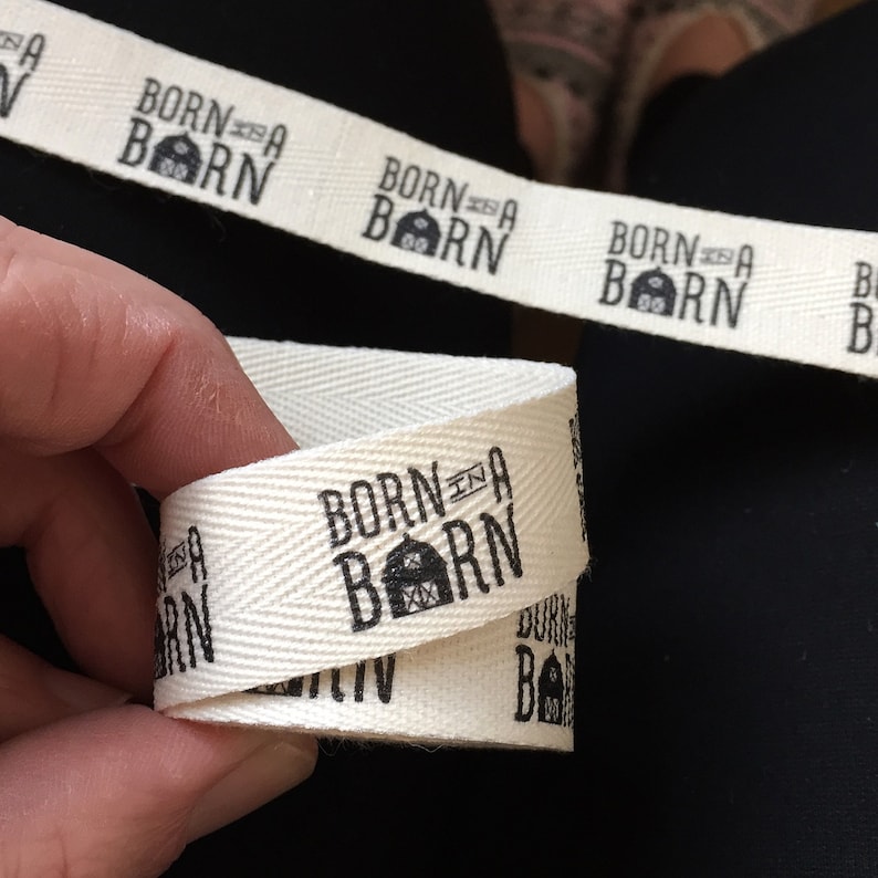 K&ouml;nnte beinhalten: Wei&szlig;es Baumwollband mit schwarzem Text, der "Born in a Barn" mit einem kleinen Scheunen-Symbol lautet. Das Band ist in der Mitte gefaltet und der Text ist auf beiden Seiten sichtbar.