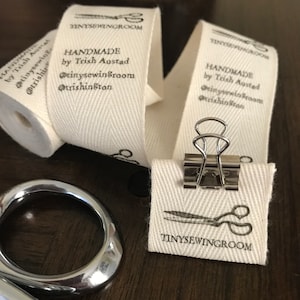 Op de afbeelding: Een close-up van een wit katoenen stoffen label met zwarte tekst die "HANDMADE by Trish Austad @tinysewingroom @trishinston TINYSEWINGROOM" luidt. Het label is bevestigd aan een rol stof met een zilveren paperclip.