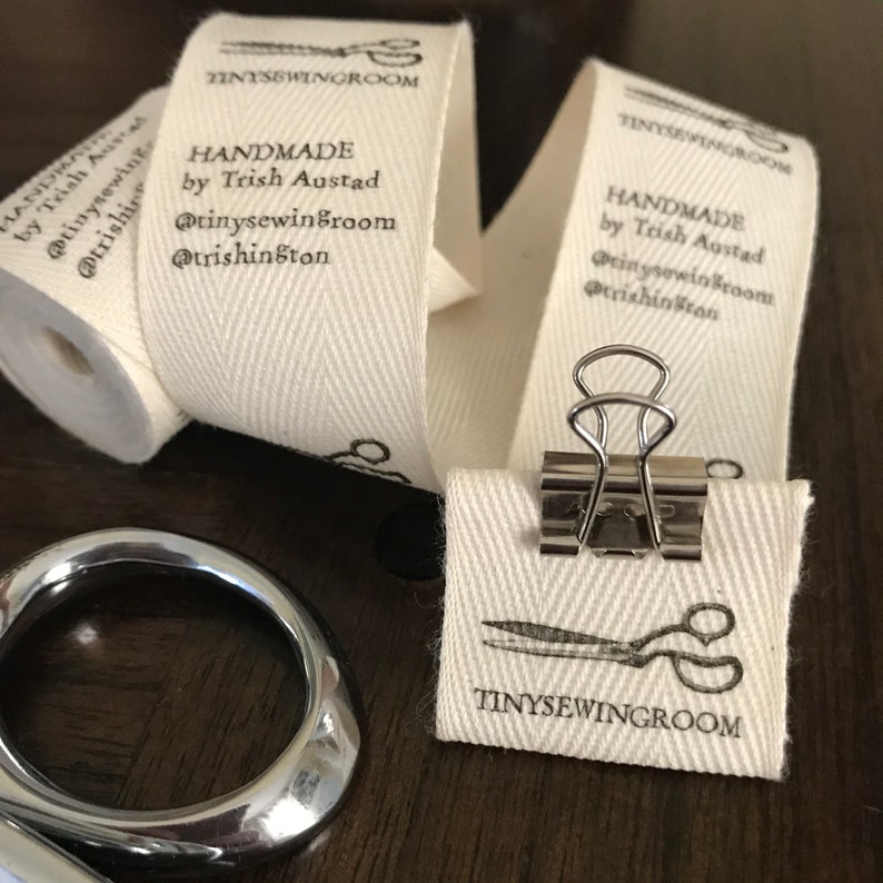 Op de afbeelding: Een close-up van een wit katoenen stoffen label met zwarte tekst die "HANDMADE by Trish Austad @tinysewingroom @trishinston TINYSEWINGROOM" luidt. Het label is bevestigd aan een rol stof met een zilveren paperclip.