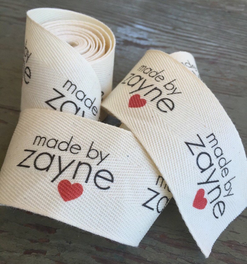 Op de afbeelding: Een rol witte katoenen stof met een zwart en rood label met de tekst "made by zayne" met een rood hart.