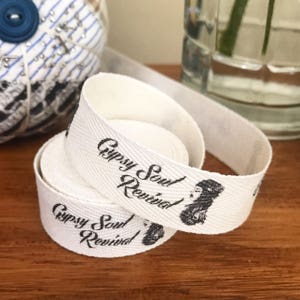 K&ouml;nnte beinhalten: Wei&szlig;es Baumwollband mit schwarzem Text, der "Gypsy Soul Revival" lautet, und einer Schwarzwei&szlig;illustration eines Frauenkopfes. Das Band ist aufgerollt und der Text ist auf beiden Seiten sichtbar.