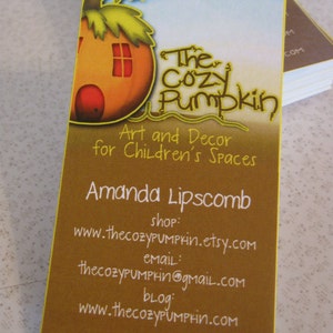 May include: Business card for The Cozy Pumpkin, a shop that sells art and decor for children's spaces. The card features a cartoon illustration of a pumpkin house with a window and a door. The text on the card reads "The Cozy Pumpkin", "Art and Decor for Children's Spaces", "Amanda Lipscomb", "shop: www.thecozypumprin.etsy.com", "email: thecozyPUMPkin@gmail.com", and "blog: www.thecozyPUMPrin.com".