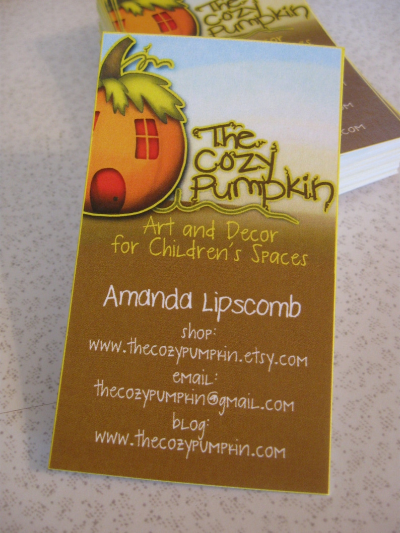 May include: Business card for The Cozy Pumpkin, a shop that sells art and decor for children's spaces. The card features a cartoon illustration of a pumpkin house with a window and a door. The text on the card reads "The Cozy Pumpkin", "Art and Decor for Children's Spaces", "Amanda Lipscomb", "shop: www.thecozypumprin.etsy.com", "email: thecozyPUMPkin@gmail.com", and "blog: www.thecozyPUMPrin.com".
