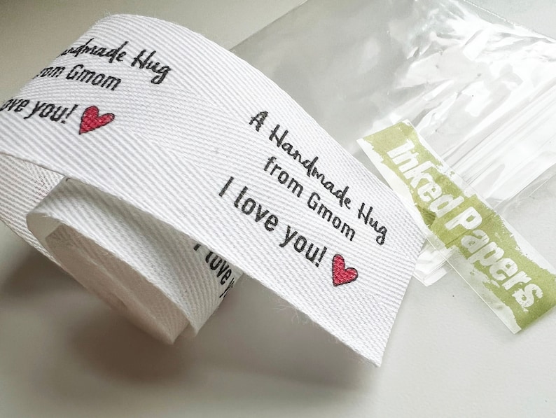 Op de afbeelding: Witte stoffen lint met de tekst "A Handmade Hug from Gmom I love you!" in zwart gedrukt met een rood hart. Het lint is opgerold en heeft een groen label met de tekst "Inked Papers" in wit gedrukt.