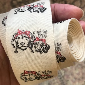 Op de afbeelding: Een wit katoenen lint met een herhalend patroon van twee honden die rode en witte stippenbandanas dragen. De tekst "Jo and the DOOD" is op het lint gedrukt.