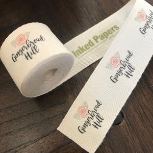 K&ouml;nnte beinhalten: Wei&szlig;es Baumwollband mit dem Text "Gingerbread Hill" und einem kleinen Blumenmuster. Das Band ist aufgerollt und hat ein gr&uuml;nes Etikett mit der Aufschrift "Inked Papers".