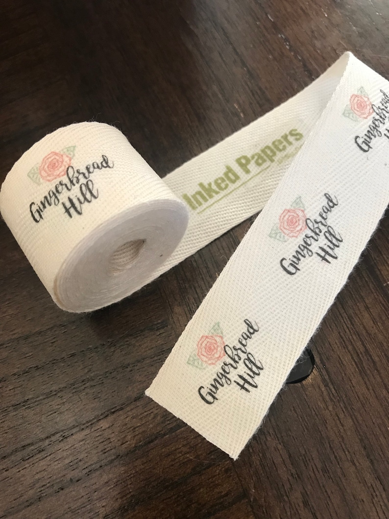 K&ouml;nnte beinhalten: Wei&szlig;es Baumwollband mit dem Text "Gingerbread Hill" und einem kleinen Blumenmuster. Das Band ist aufgerollt und hat ein gr&uuml;nes Etikett mit der Aufschrift "Inked Papers".