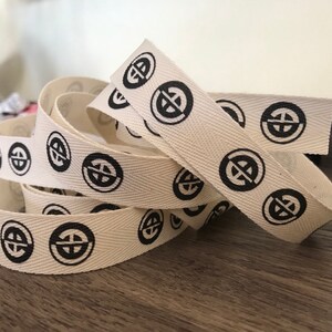 K&ouml;nnte beinhalten: Nahaufnahme eines wei&szlig;en Baumwoll-Twillbandes mit einem sich wiederholenden schwarzen Muster aus einem Kreis mit einem Kreuz darin. Das Band ist auf einer Holzoberfl&auml;che gefaltet und gestapelt.